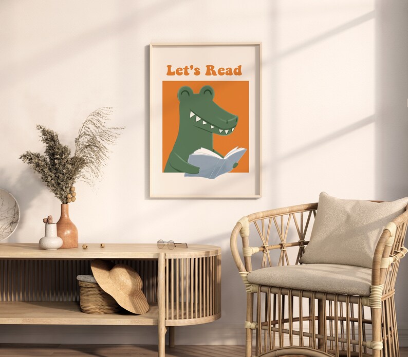 A framed "Let's Read" poster featuring a green alligator reading a book against an orange background, hanging in a cozy, sunlit reading corner with a wicker chair and a wooden console table.