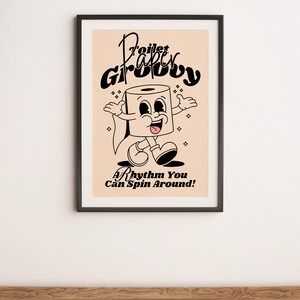Op de afbeelding: Een zwart ingelijste poster met een licht perzikkleurige achtergrond. De poster toont een cartoon toiletpapierrol met een glimlachend gezicht en de tekst "Toilet Paper Groovy" in een retro-lettertype. De tekst "A Rhythm You Can Spin Around!" staat onder de cartoon.
