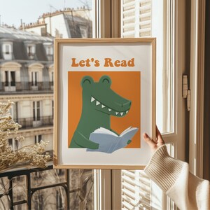 A hand holds a framed "Let's Read" poster with a green alligator reading a book against an orange background, in front of a Parisian balcony backdrop.