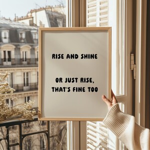 May include: A framed print with the text "RISE AND SHINE OR JUST RISE, THAT'S FINE TOO" in black font. The print is held near a window with a view of a building with multiple windows. The frame is made of light-colored wood.