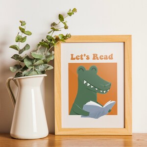 A framed "Let's Read" poster with a green alligator reading a book against an orange background, displayed on a wooden shelf next to a white vase with green foliage.