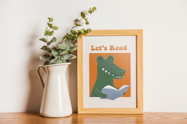 A framed "Let's Read" poster with a green alligator reading a book against an orange background, displayed on a wooden shelf next to a white vase with green foliage.