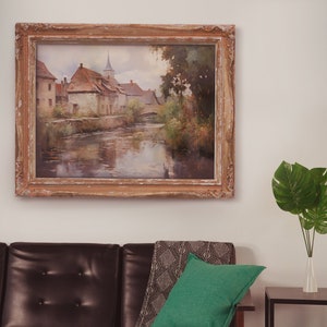 Classically framed artwork of an old European village by a calm river, hung above a leather sofa with greenery in the room.