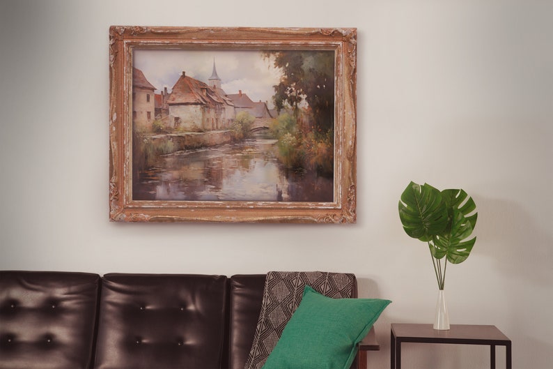 Classically framed artwork of an old European village by a calm river, hung above a leather sofa with greenery in the room.