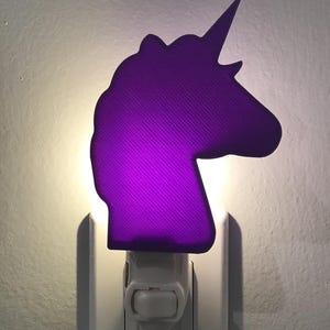 May include: A purple unicorn-shaped night light plugged into an electrical outlet. The light is on and illuminating the room.