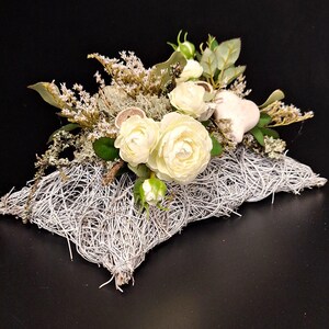 Könnte beinhalten: Dekorative Blumengesteck mit weißen Rosen, grünen Blättern und getrocknetem Laub. Die Blumen sind auf einer weißen, gewebten Basis angeordnet, mit einem herzförmigen Akzent. Der Hintergrund ist schwarz.