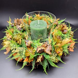 Könnte beinhalten: Ein dekoratives Centerpiece mit einer grünen Säulenkerze in einer durchsichtigen Glasschale, umgeben von einem Kranz aus künstlichem Grün, Blättern und kleinen braunen Akzenten. Die Anordnung befindet sich vor einem dunklen Hintergrund.