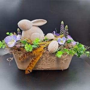 Könnte beinhalten: Ein grauer Betontopf mit dem Text "Jardin de Fleurs" auf der Vorderseite. Der Topf ist gefüllt mit grünem Moos, künstlichem Grün, lila Blumen und einem weißen Keramik-Hasen. Es befinden sich auch weiße Keramik-Eier im Topf.