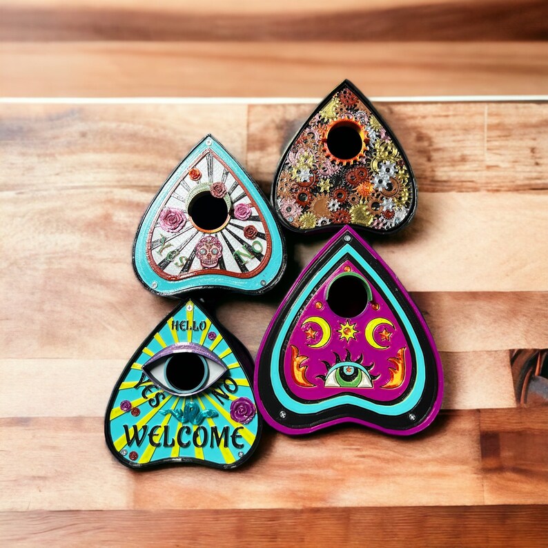 May include: Four wooden planchette boards with different designs. The planchette boards are shaped like a heart with a pointed top. The designs include a skull and crossbones, a colorful eye, a gear pattern, and a crescent moon and stars.