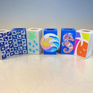 May include: A collection of six decorative tissue box covers. The boxes feature various designs, including sea creatures, a sunset, and a seahorse. The color palette includes blue, white, orange, and yellow. The boxes are rectangular with a square opening at the top.