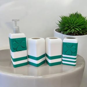 May include: Set of four white bathroom accessories with green clover accents and green and white stripes. The set includes a soap dispenser, a toothbrush holder, and two tumblers.