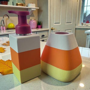 May include: A decorative soap dispenser and a matching container, both designed in the style of candy corn. The dispenser has a burgundy pump and a white, orange, and yellow color scheme. The container is a similar shape and color.