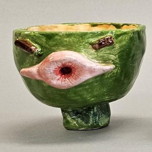 Puede incluir: Una fila de cinco macetas de cerámica con forma de cara, caprichosas y de varios colores. Las macetas son verdes, azules, rosas, moradas y amarillas, cada una con rasgos faciales y expresiones únicas. Tienen un aspecto artesanal y artístico.