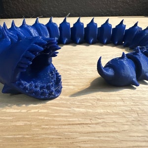 May include: A blue, segmented, 3D-printed articulated creature with a large open mouth and a curved horn. The creature is made of plastic and has a spiky dorsal fin. The design is reminiscent of a sea serpent.