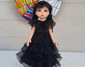 May include: A doll with black pigtails, dressed in a black, layered dress. The doll is positioned in front of a circular artwork, half colourful and half a spiderweb design. The background is a light wooden wall.