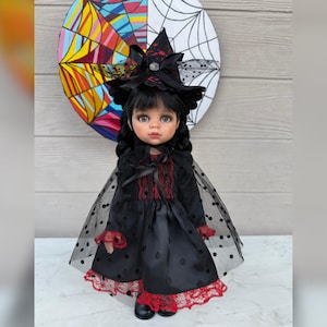 May include: A doll dressed in a black witch costume with a matching hat, cape, and dress. The dress has red lace trim and black polka dots. The doll has black hair and is wearing black shoes. A colorful abstract art piece is in the background.