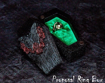 May include: Black wooden coffin-shaped ring box with a red and black butterfly design and a green velvet interior. The box is open and a gold ring is inside. "Proposal Ring Box" is written in white text below the box.