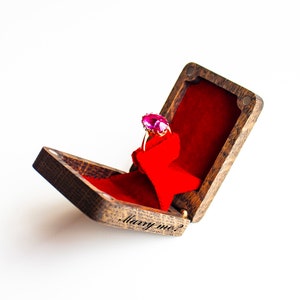 May include: A wooden ring box with a red velvet interior. The box is open and a ring with a pink gemstone is inside. The box is engraved with the words "Marry me?"