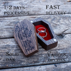 May include: A wooden coffin-shaped ring box with a red velvet interior, holding two silver rings. The box is open, revealing the rings. The lid is engraved with a skeleton couple and the text "The Death Do Us Part". The box is made of solid oak.