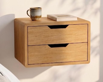 Può includere: Un comodino sospeso in legno color marrone chiaro con due cassetti, dotato di maniglie integrate. Il comodino è montato a parete e sostiene una tazza in ceramica e un libro chiuso sulla superficie superiore, mostrando una finitura a venature naturali.