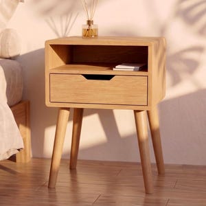 Peut inclure: Une table de chevet en bois clair dotée d'un tiroir unique avec une poignée encastrée et d'une étagère de rangement ouverte au-dessus. Le meuble repose sur quatre pieds fuselés et présente des coins arrondis avec un diffuseur en verre.