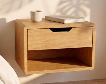 May include: A light-toned wooden floating nightstand featuring a single drawer with a recessed handle and an open storage shelf below. A small ceramic cup and a closed book are placed on the flat top surface of the wall-mounted bedside unit.
