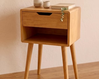 Könnte beinhalten: Ein Nachttisch aus hellem Holz mit vier konisch zulaufenden Beinen, einer Schublade mit eingelassenem Griff und einem offenen Ablagefach. Obenauf stehen eine kleine Keramikvase, eine passende Tasse, ein geschlossenes Buch und ein Eukalyptuszweig.