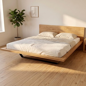 Op de afbeelding: Een laag platformbedframe gemaakt van lichtgekleurd hout met een opvallend hoofdeinde. Het bed heeft een zwarte metalen onderstel en is opgemaakt met een wit matras, twee bijpassende kussens en een lichtgekleurde sprei.