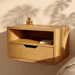 May include: A light oak floating nightstand featuring a rectangular open shelf and a single drawer with a recessed handle. The wooden unit is mounted to a wall and holds a small glass reed diffuser with wooden sticks and a white rectangular object.