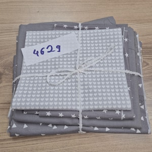 Könnte beinhalten: Ein Bündel grauer Stoffquadrate, die mit weißer Schnur zusammengebunden sind. Der Stoff weist weiße Herz-, Stern- und Dreieckmuster auf. Ein weißes Etikett mit der Nummer "4629" ist angebracht. Der Stoff ist auf einer hellen Holzoberfläche gestapelt.