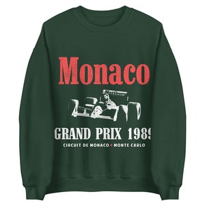May include: A dark green sweatshirt with a white vintage-style graphic of a race car driver in a Formula One car. The text "Monaco Grand Prix 1989" is printed below the car, with the words "Circuit de Monaco Monte Carlo" printed below the text.