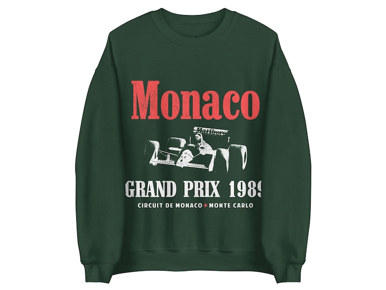 May include: A dark green sweatshirt with a white vintage-style graphic of a race car driver in a Formula One car. The text "Monaco Grand Prix 1989" is printed below the car, with the words "Circuit de Monaco Monte Carlo" printed below the text.