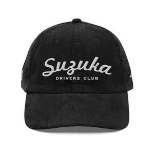 Puede incluir: Gorra de béisbol de pana negra con el texto bordado en blanco "Suzuka Drivers Club" en cursiva. La gorra tiene una visera curva y un botón en la parte superior. Un pequeño logotipo blanco está en el lateral.
