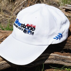 May include: White baseball cap with an embroidered logo that says "Silverstone British Grand Prix" with a blue and red race car. The cap has a blue embroidered leaf on the side.