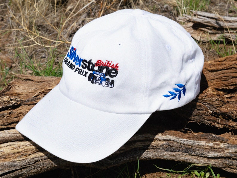 May include: White baseball cap with an embroidered logo that says "Silverstone British Grand Prix" with a blue and red race car. The cap has a blue embroidered leaf on the side.