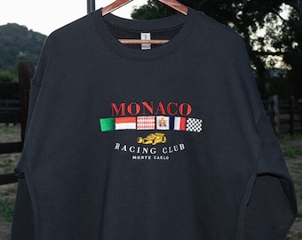 May include: Black sweatshirt with red embroidered text that reads "MONACO RACING CLUB MONTE CARLO". The text is surrounded by small embroidered flags of Italy, Indonesia, Monaco, Great Britain, France, and a checkered flag.