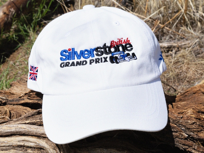 May include: White baseball cap with an embroidered logo that says "Silverstone British Grand Prix" with a red, blue, and black color scheme. The logo also includes a small image of a race car. The cap has a small embroidered Union Jack on the side.