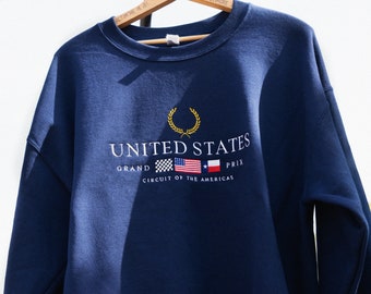 May include: A navy blue sweatshirt with a white embroidered logo that says "UNITED STATES GRAND PRIX CIRCUIT OF THE AMERICAS". The logo also includes a laurel wreath, a checkered flag, and the American and Texas flags.