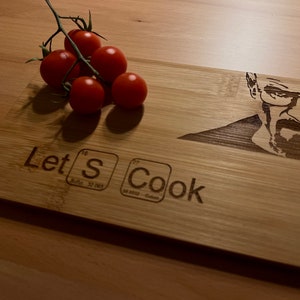May include: A wooden chopping board with a burnt-in image of a man with a goatee and glasses. The board also has the text "Let's Cook" with the letter "S" replaced by the chemical symbol for sulfur. There are five red cherry tomatoes on a vine on the top left of the board.