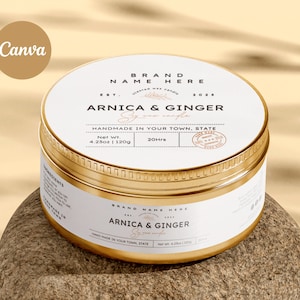 May include: A gold metal candle jar with a white label. The label says "BRAND NAME HERE" and "ARNICA & GINGER Soy wax candle". The jar is sitting on a light brown surface.