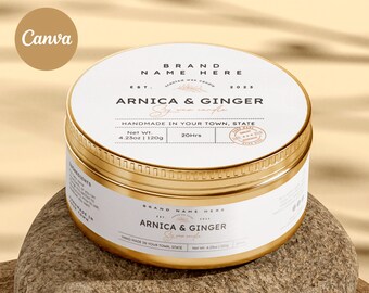 Może przedstawiać: Złoty metalowy słoik na świecę z białą etykietą. Etykieta zawiera napis "BRAND NAME HERE" i "ARNICA & GINGER Świeca sojowa". Słoik stoi na jasnobrązowej powierzchni.