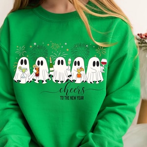 K&ouml;nnte beinhalten: Gr&uuml;nes Sweatshirt mit einer Reihe von Cartoon-Geistern, die Cocktails, Champagner und Wein halten. Die wei&szlig;en Geister mit schwarzen Augen stehen vor einem Feuerwerk. Der Text "cheers TO THE NEW YEAR" ist in einer Schreibschrift geschrieben.