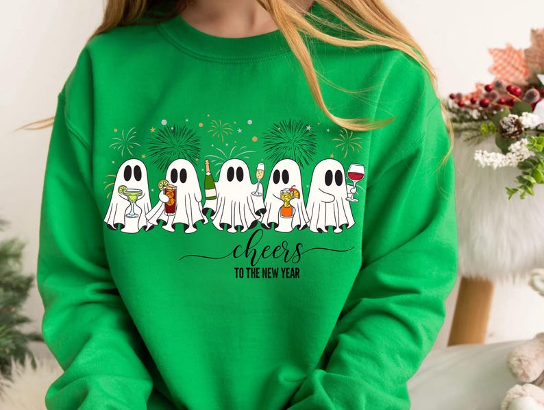 K&ouml;nnte beinhalten: Gr&uuml;nes Sweatshirt mit einer Reihe von Cartoon-Geistern, die Cocktails, Champagner und Wein halten. Die wei&szlig;en Geister mit schwarzen Augen stehen vor einem Feuerwerk. Der Text "cheers TO THE NEW YEAR" ist in einer Schreibschrift geschrieben.