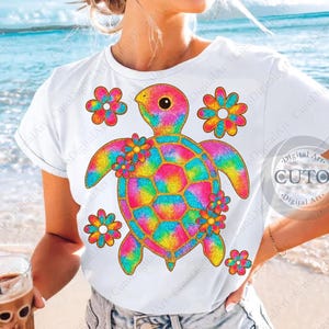 以下が含まれることがあります： カラフルなウミガメのデザインが施された白いTシャツ。カメは虹色のタイダイ模様と金色のグリッターのディテールがあり、カラフルな花に囲まれています。カジュアルなビーチをテーマにしたデザインです。