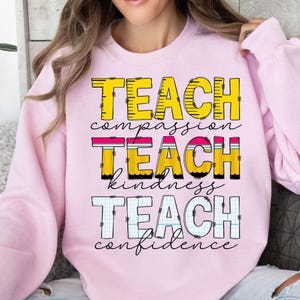 Puede incluir: Sudadera rosa con el texto "Enseñar compasión, Enseñar amabilidad, Enseñar confianza" en letras amarillas, rosas y blancas con un diseño de lápiz.