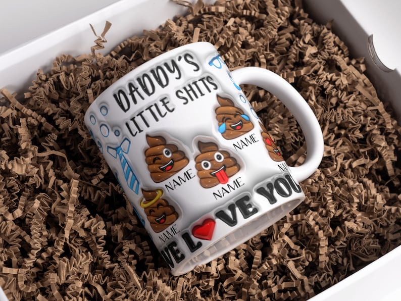 K&ouml;nnte beinhalten: Eine wei&szlig;e Keramiktasse mit dem Text "Daddy's Little Shits" und einem roten Herzen. Die Tasse hat eine blaue Krawatte und mehrere Kack-Emojis mit verschiedenen Gesichtsausdr&uuml;cken. Der Text "We Love You" ist um den Boden der Tasse geschrieben.