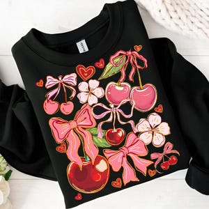 May include: Black sweatshirt with a colorful print of cherries, bows, and flowers. The print features pink, red, and gold accents.
