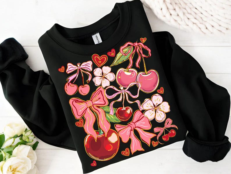May include: Black sweatshirt with a colorful print of cherries, bows, and flowers. The print features pink, red, and gold accents.