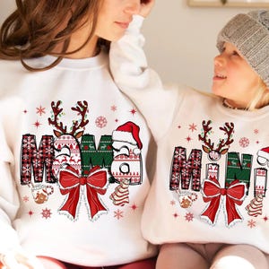 May include: White sweatshirts with a Christmas-themed design. The word "MAMA" is displayed with reindeer antlers, a Santa hat, and a red bow. The sweatshirts are decorated with snowflakes and gingerbread cookies.