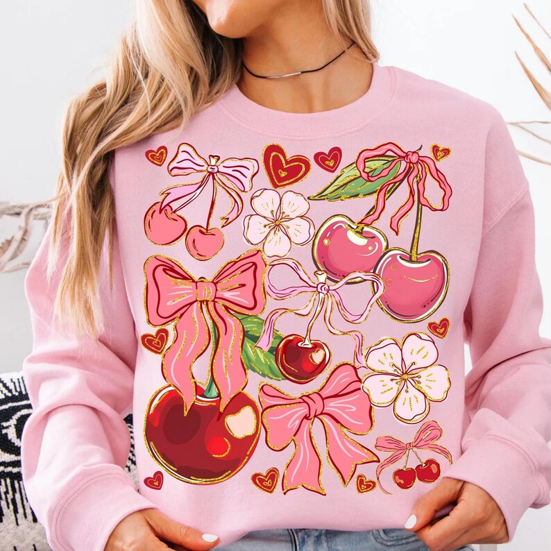 May include: Pink sweatshirt with a graphic print of cherries, bows, and hearts. The cherries are red and have green stems and leaves. The bows are pink and have gold glitter. The hearts are red and have a white outline.